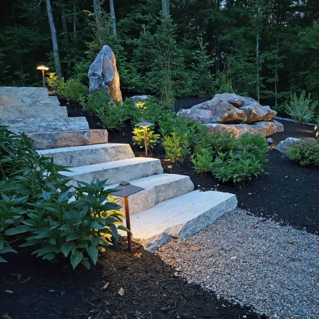 Nighttime garden scene with stone steps, large rocks, greenery, small trees, and landscape lighting.