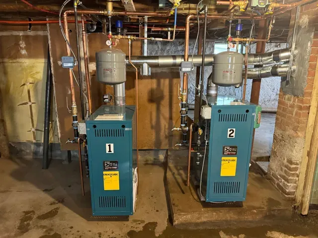 Two blue industrial boilers in a basement utility room with pipes and brick walls.