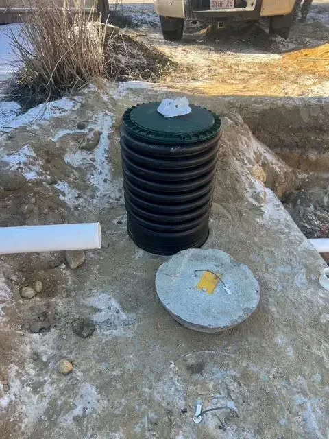 Black ribbed utility pipe in concrete pit, with two round metal lids nearby and a truck in the background