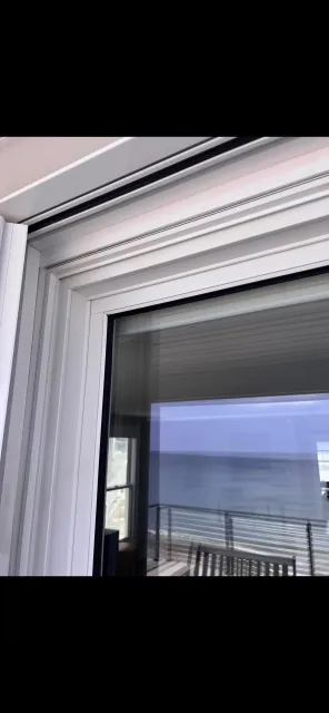 Close-up of a white sliding glass door frame with an outdoor balcony visible through the glass, showing a railing and a distant ocean view.