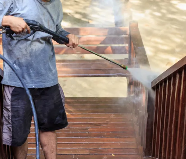 A person using a pressure washer to clean wooden stairs and railings outdoors, with water spraying and mist visible. The person is wearing a gray shirt and black shorts.