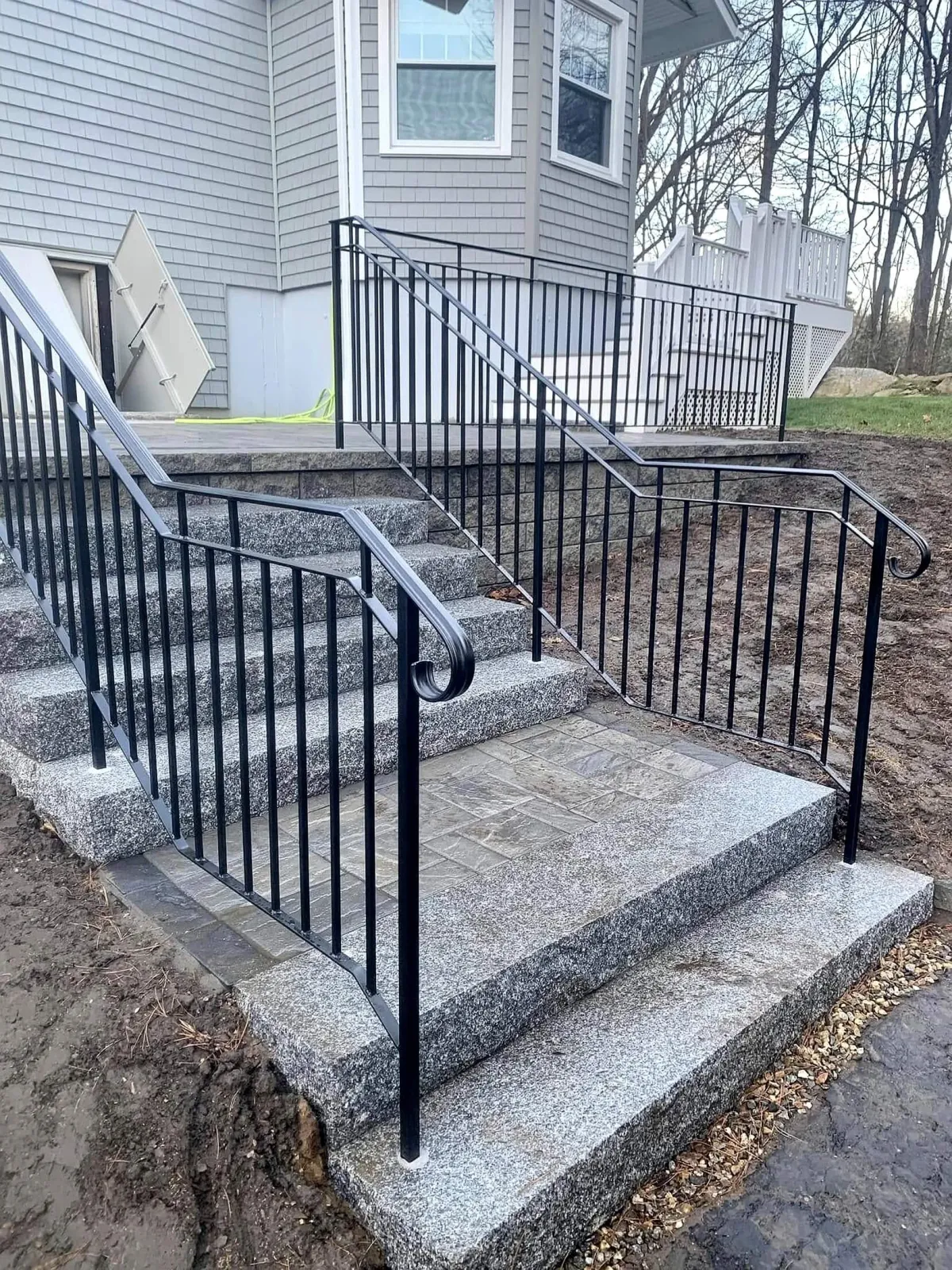 Concrete steps with black metal railing leading up to a house with grey siding, white window frames, and a small deck in the backyard, surrounded by dirt and some grass and trees in the background.