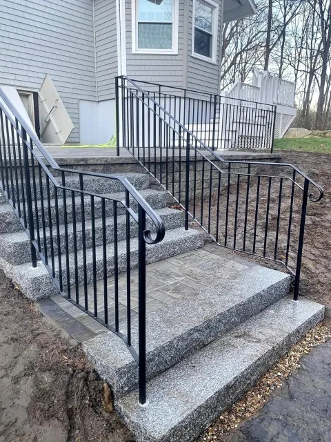 Concrete steps with black metal railing leading up to a house with grey siding, white window frames, and a small deck in the backyard, surrounded by dirt and some grass and trees in the background.