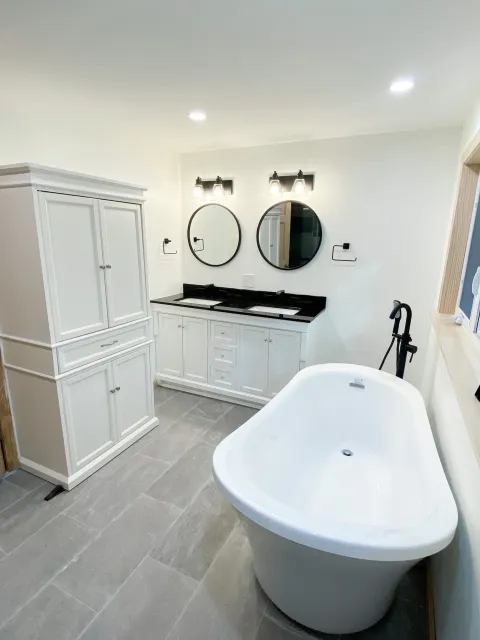 A modern bathroom with a white freestanding bathtub, white vanity with two sinks, two round mirrors, black fixtures, and gray floor tiles.