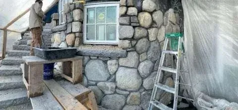 Stone masonry work in progress on a building facade with a window, ladder, and construction tools.