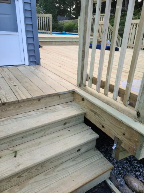 Newly built wooden deck with steps and railing, partially completed, with a pool visible in the background.