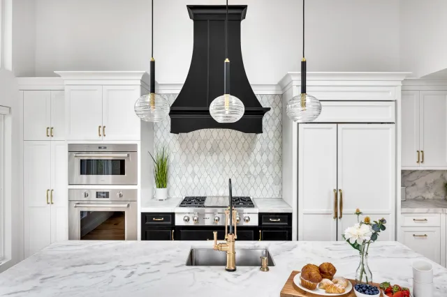 Modern kitchen with white cabinets, black stove and range hood, marble island with gold faucet, pendant lights, and a backsplash with a geometric pattern. Countertop has a potted plant, muffins, flowers, berries, and strawberries.