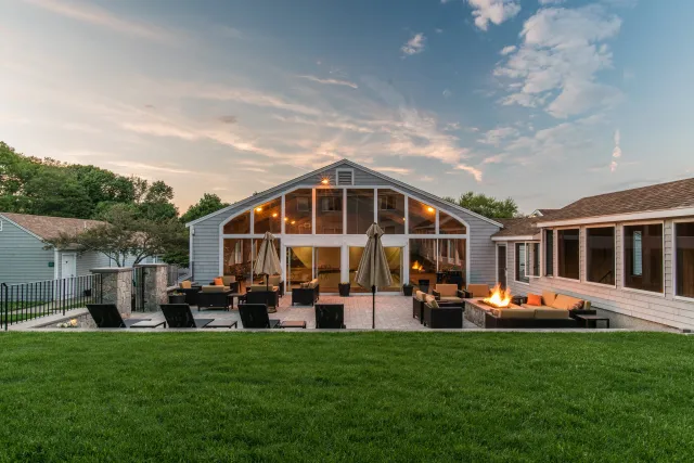 A spacious backyard patio with outdoor furniture, umbrellas, and a fire pit, adjacent to a house with large windows and a glass room under a partly cloudy sky at sunset.
