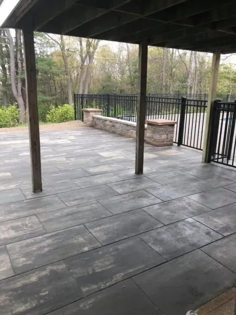 Covered stone patio with dark posts, a built-in bench, and black railing overlooking trees and shrubs