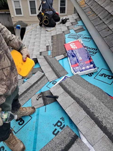 People removing torn blue and gray roof shingles with tools on a sloped rooftop.