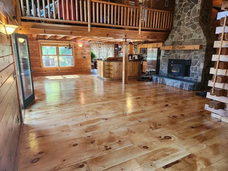 Spacious living area in a log cabin with honey-colored wooden floors, wood-paneled walls, a stone fireplace, and a kitchen area with a breakfast bar and stainless steel appliances.