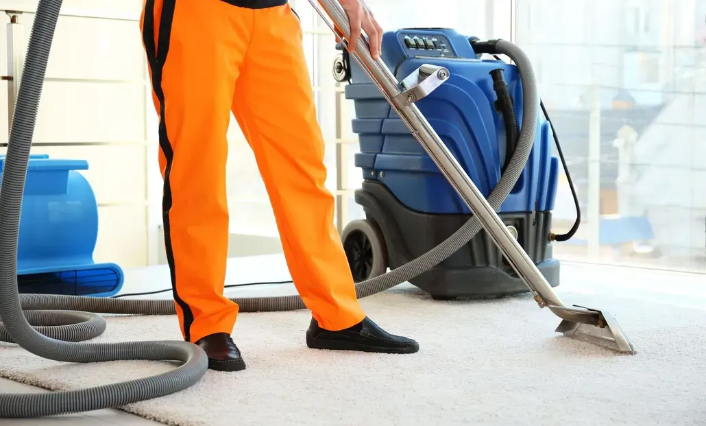 Worker in orange pants vacuuming a carpet with a blue industrial cleaner.