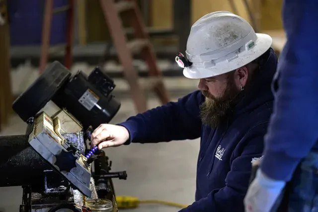 Construction worker in a hard hat, using a tool on machinery indoors.