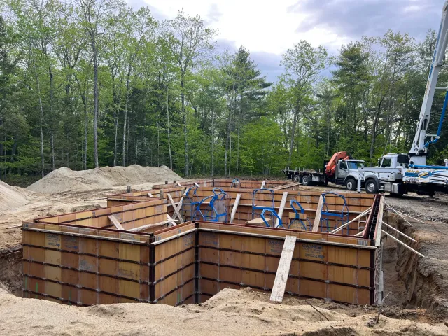 Construction site with foundation formwork and blue support braces, surrounded by dirt and sand, with trees and cloudy sky in the background. Construction trucks and equipment are present.