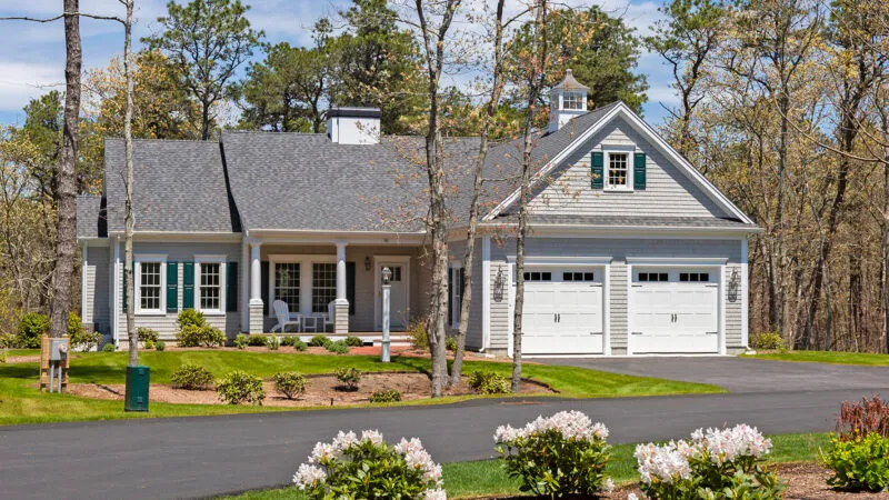 Gray single-story house with porch, green shutters, flower beds, and trees – McPhee Associates of Cape Cod.