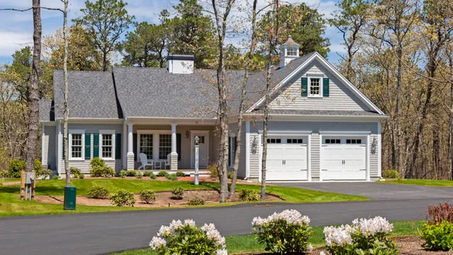 Gray single-story house with porch, green shutters, flower beds, and trees – McPhee Associates of Cape Cod.