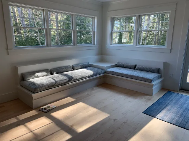 Bright living room with wood floors, large windows, built-in window seat with gray cushions, and a small blue rug near the door.