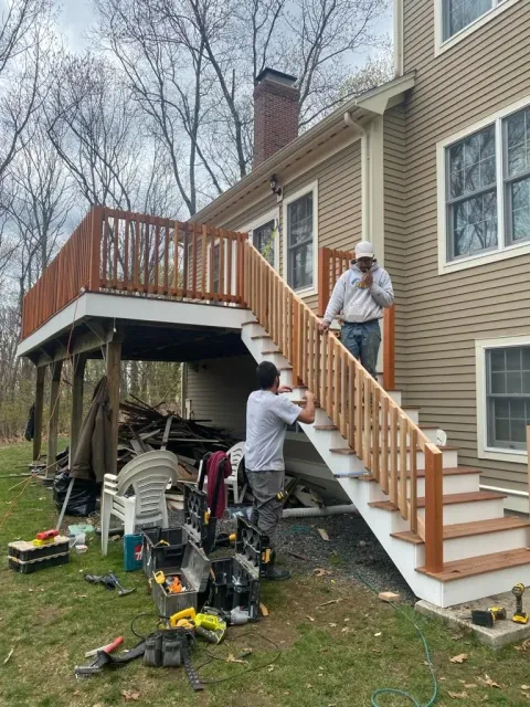 Two construction workers assemble a wooden staircase leading to an elevated deck on the side of a tan house.