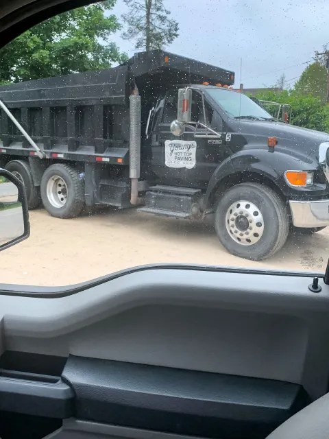 A black dump truck with a sign on the side reading 'Young's Hot Top Paving' parked on a dirt surface, viewed through a vehicle window with raindrops.