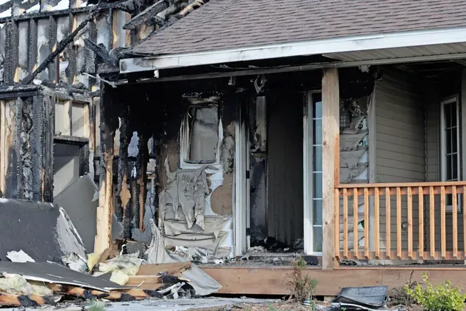 Severely burned house exterior with blackened walls, debris, and a damaged porch.