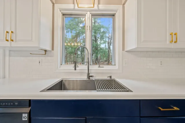 A stainless steel kitchen sink with a gooseneck faucet under a window, set in white countertops between dark blue cabinets.