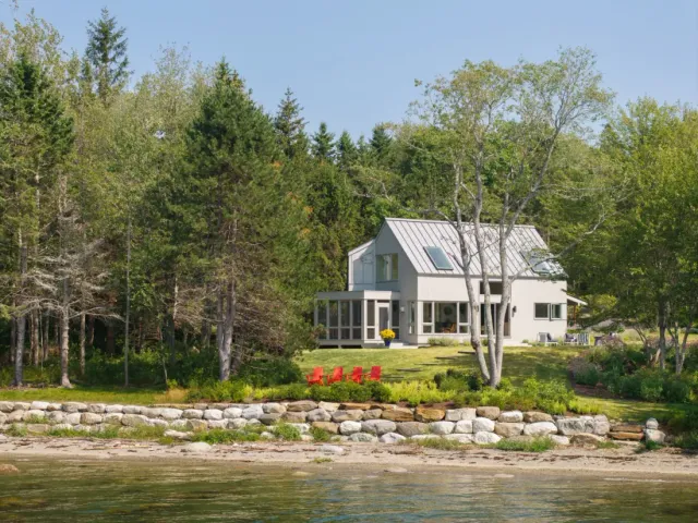 A modern house by the water, designed by a Maine architect, features large windows and four red chairs on the lawn facing the tree-lined shoreline.