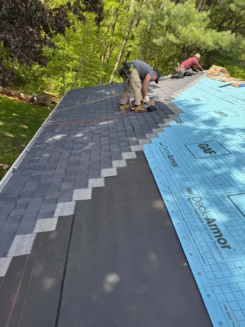 Workers installing roof shingles beside blue underlayment on a sloped roof.