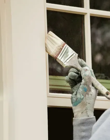 Hand wearing a paint-splattered glove painting a cream window frame with a brush