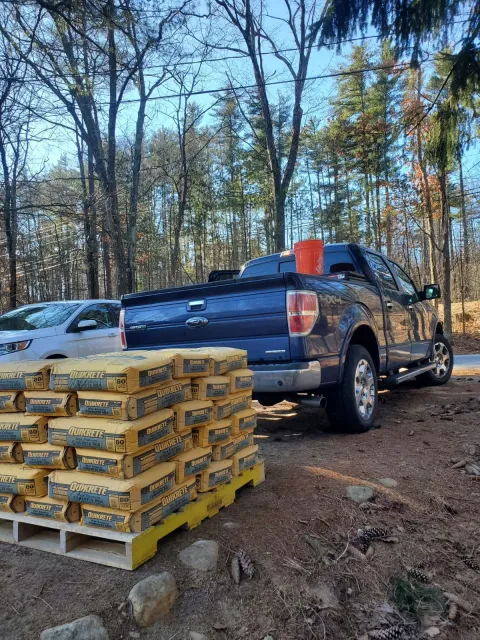 Blue pickup truck parked near stacked yellow bags on a pallet; a bucket is in the bed.