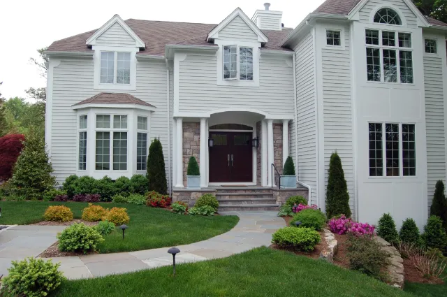 Front view of a large, white, two-story house with multiple windows, a brown door, and a landscaped lawn with bushes, flowers, and pathway.