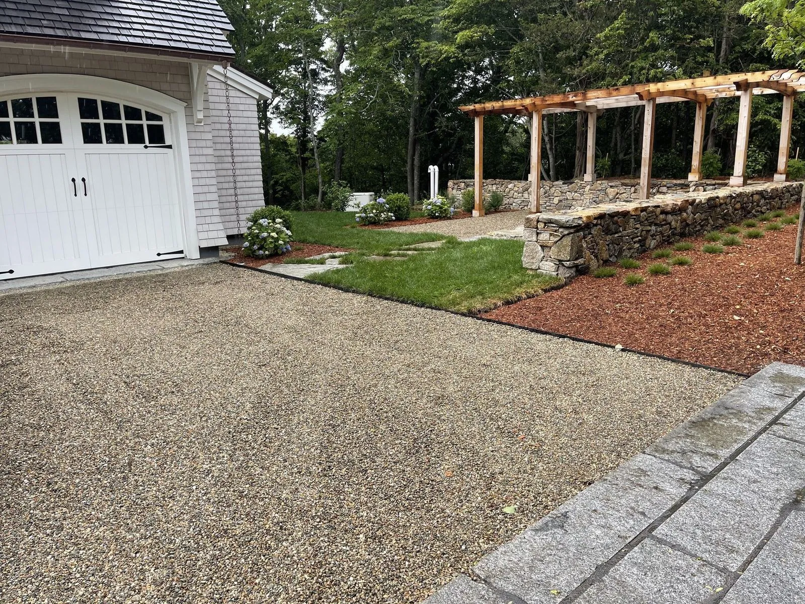 A custom timber-and-stone pergola anchoring a pea-gravel drive court at a Cape Cod home, with a hand-set fieldstone retaining wall behind