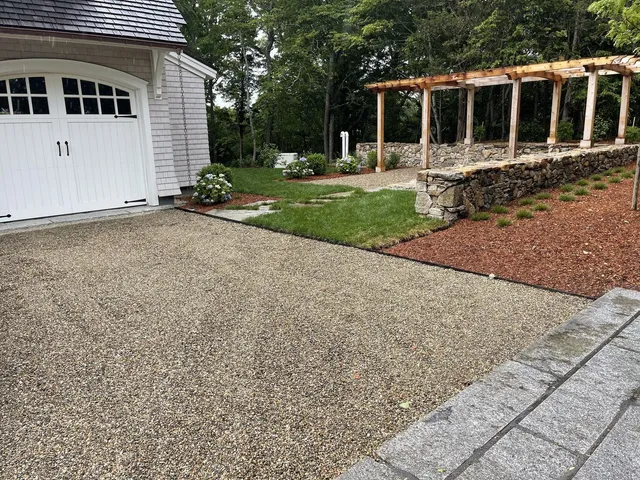 A custom timber-and-stone pergola anchoring a pea-gravel drive court at a Cape Cod home, with a hand-set fieldstone retaining wall behind