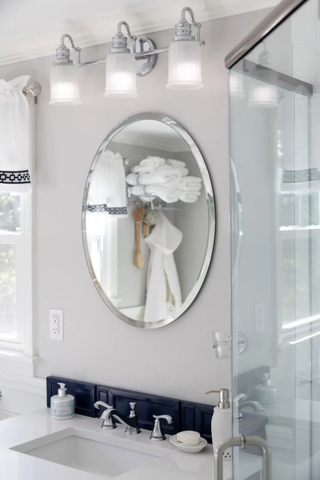 A modern bathroom vanity with a white countertop, dark blue tiled backsplash, oval mirror, and three glass pendant lights.