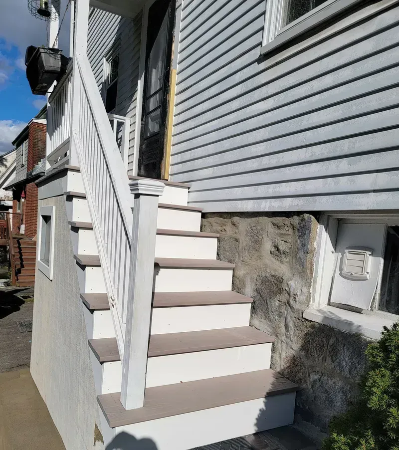 Exterior staircase leading up to a house entrance. White railing and stairs, gray siding, stone foundation.