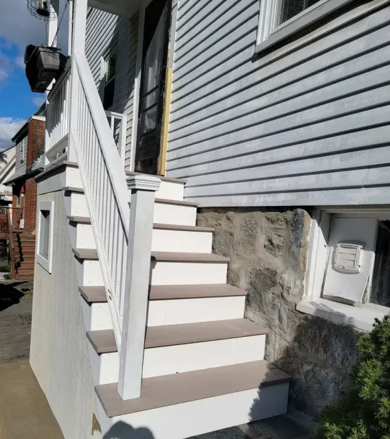 Exterior staircase leading up to a house entrance. White railing and stairs, gray siding, stone foundation.