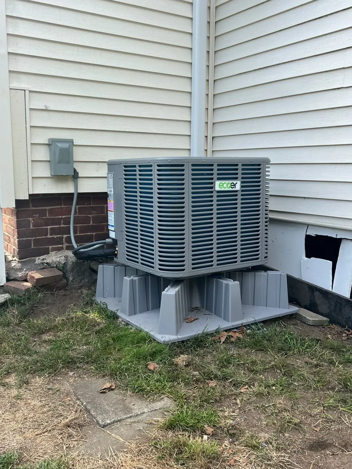 Outdoor HVAC unit on foam pad beside a house with beige siding and brick foundation.