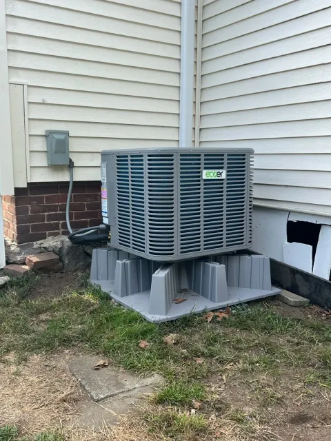 Outdoor HVAC unit on foam pad beside a house with beige siding and brick foundation.