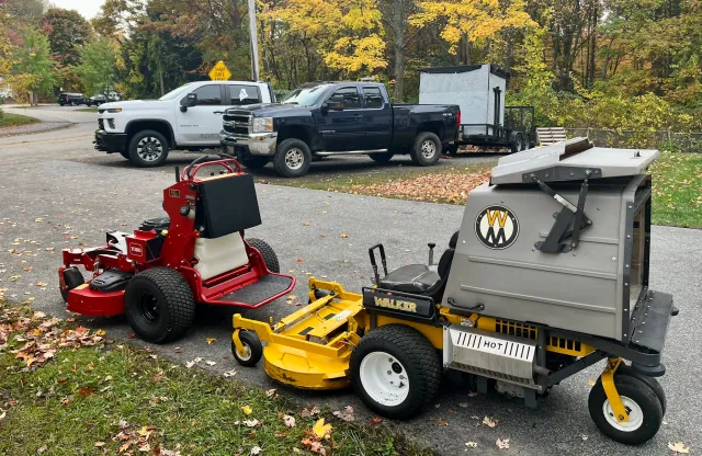 Two large trucks parked on a paved area with trees and autumn leaves in the background, and two ride-on lawn mowing machines in the foreground.