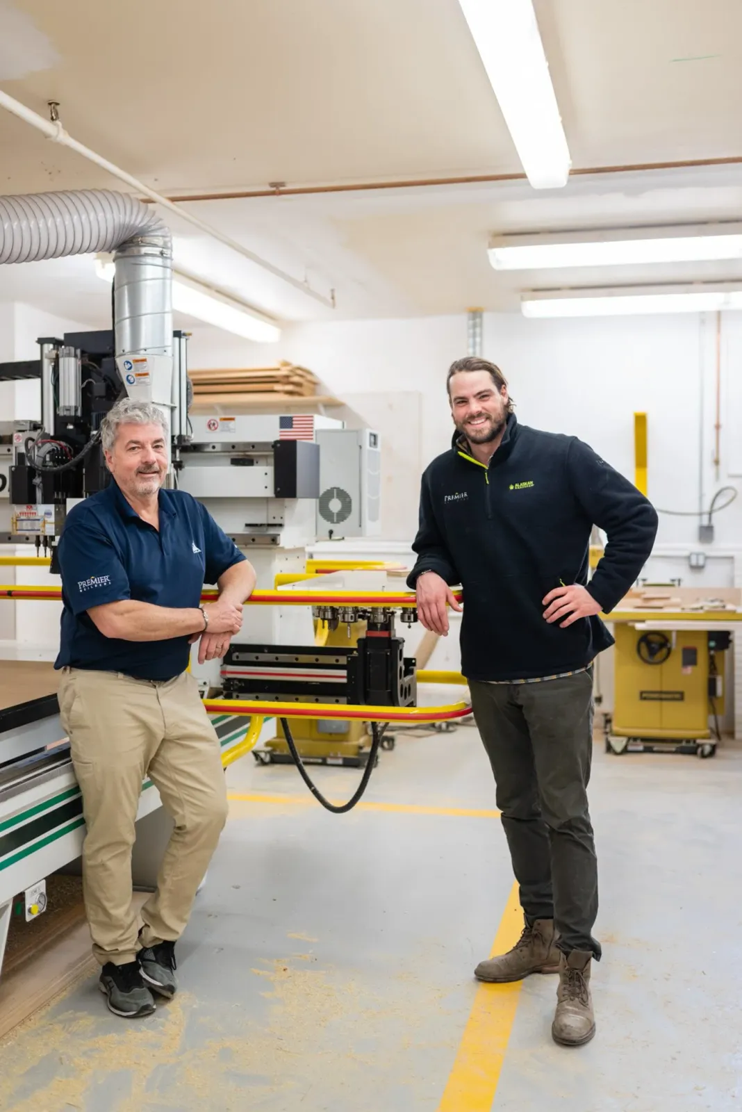 Two men standing in a woodworking shop, smiling, with woodworking equipment and wood in the background.