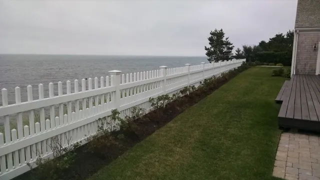A white picket fence lines a green lawn overlooking a calm, gray sea under an overcast sky.