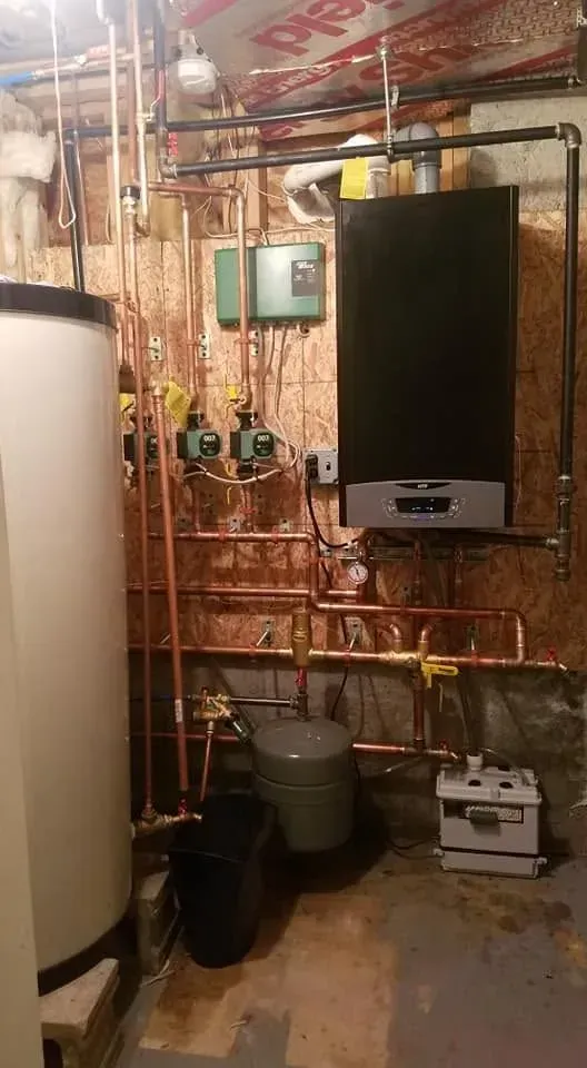 Utility room with boiler, copper pipes, water tank, and wall-mounted control panel on unfinished wall.