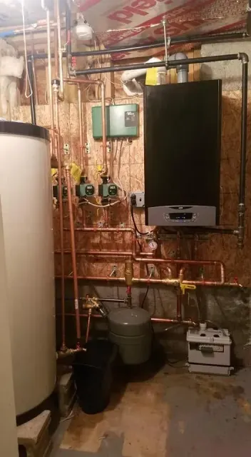Utility room with boiler, copper pipes, water tank, and wall-mounted control panel on unfinished wall.