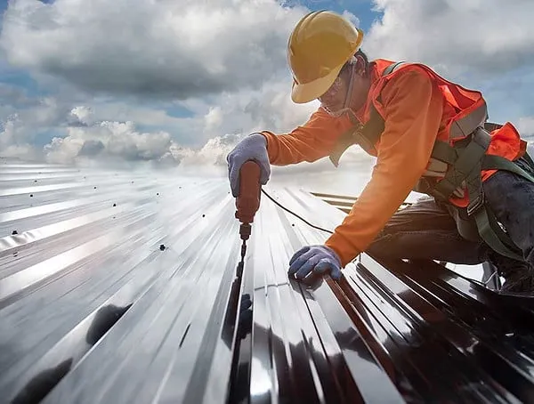 Construction worker using a power tool to secure a metal panel during industrial roof repairs under cloudy skies in Rhode Island.