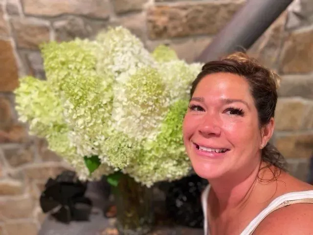 A woman is smiling in front of a vase of flowers.