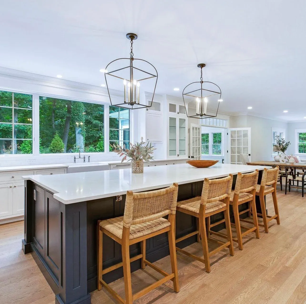Modern kitchen with a large white island, wooden stools, pendant lights, large windows showing greenery outside, and a dining area in the background.