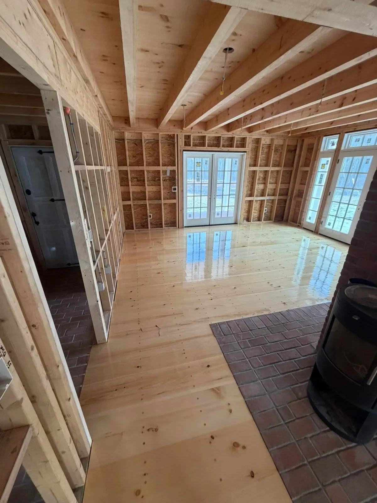 Interior of a house under construction with unfinished wooden walls, exposed ceiling beams, French doors, and a wood stove on a brick hearth.