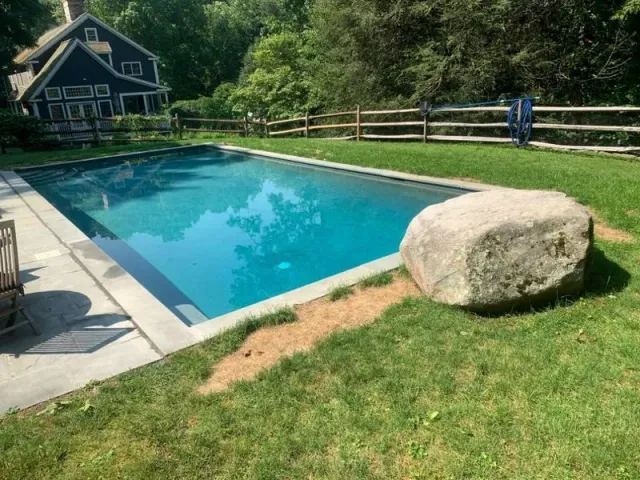 A bright blue rectangular swimming pool with a stone patio edge and a large boulder sitting on the grass beside it.