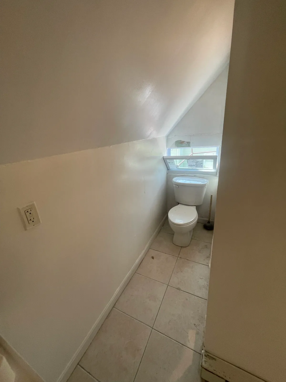 A small bathroom with tiled floor, a white toilet, a window with a screen, and a toilet brush beside it.