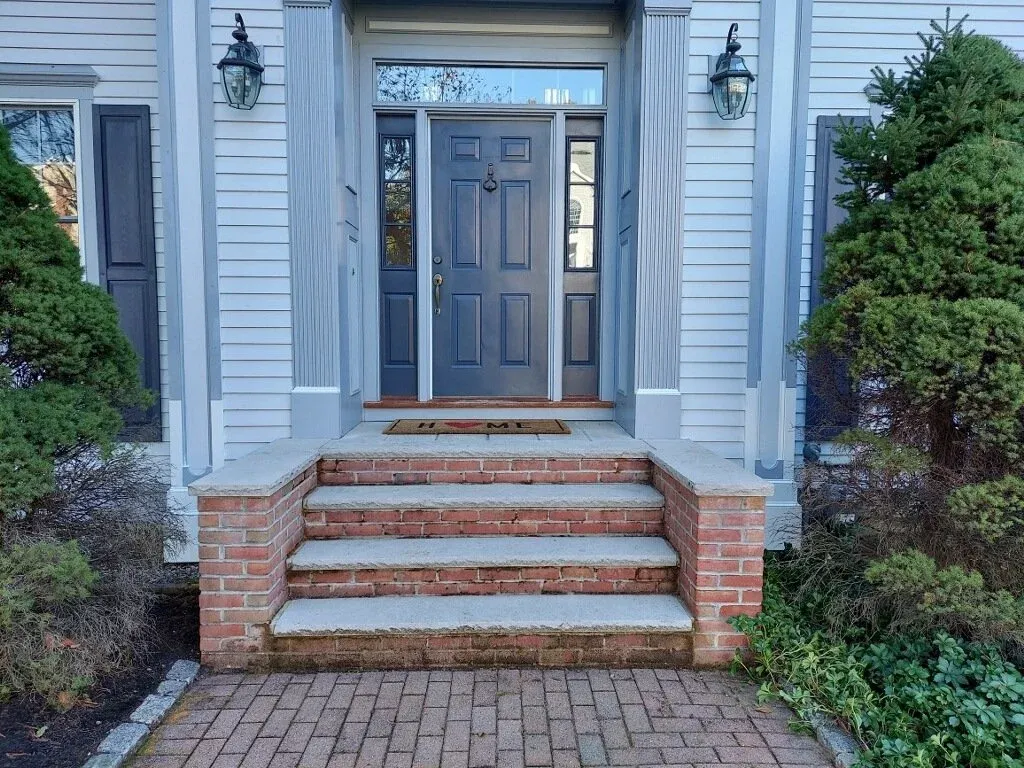 Front entrance of a house with a dark blue door, brick steps, outdoor lantern lights, and greenery on both sides.