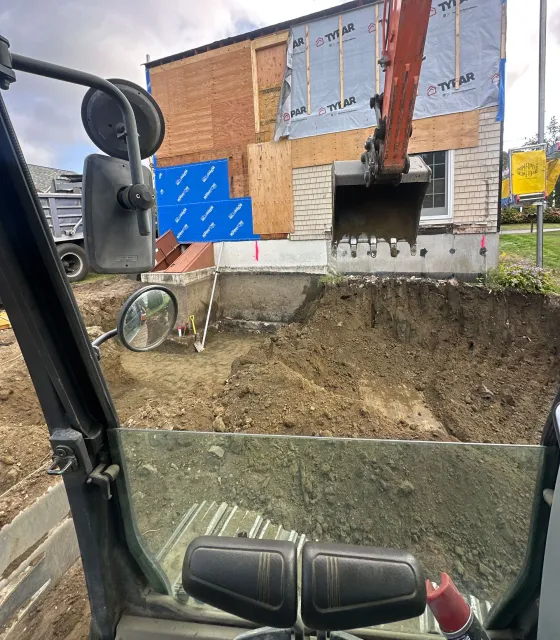 View from inside an excavator cab showing its bucket digging in front of a construction site with exposed home framing.